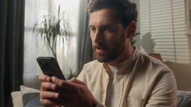 Düşünceli rüyalı beyaz erkek ev sahibi serbest kiracı evinde cep telefonu uygulaması kullanıyor. Apartman dairesinde internetten alışveriş yapıyor. Kanepede rahat ol.