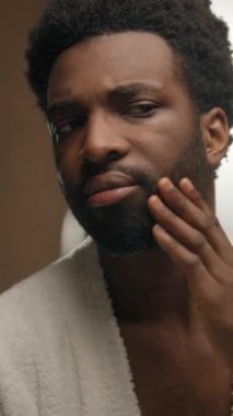 Aynaya bakan Afrikalı Amerikalı adam otel banyosunda gülümseyen sakallı adam sakal jölesi tıraş eden yumuşak cilt tıraşı sabah temizliği yapan erkek kozmetik ürünleri nemlendirici.