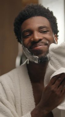 Afro-Amerikalı adam sakal tıraşı köpüklü jeli banyoda havluyla siliyor gülümseyen erkek yüzü traş oluyor ayna kozmetik ürününe bakıyor pürüzsüz cilt hijyenine bakıyor