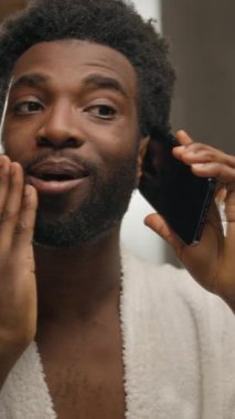 Aynada yansıyan banyoya bakan Afrikalı bir adam cep telefonuyla konuşan, yüz bakımına dokunan, sakallı, sakallı, erkek, otel banyosunda akıllı telefon görüşmesi yapan, sakallı, sakallı bir adam.