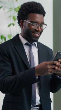 Ofiste akıllı telefon kullanan gülümseyen Afro-Amerikalı işadamı cep telefonlu neşeli profesyonel girişimciye bakan etnik erkek adam internet başarısına göz atan mesajları kontrol ediyor