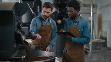 Çok ırklı Kafkas Afrikalı baristler kahve kızartma makinesinin işlevselliğini test ediyorlar. Fasulye aroma ekipman kontrol sürecini denetliyorlar. Endüstriyel espresso makinesini kızartıyorlar. İki teknisyen kontrol listesi panosunu öğütüyorlar.