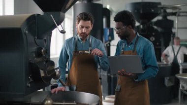 Çok ırklı kahve fabrikası ekibi online laptop teknolojili espresso aroması kullanarak kızarma verilerini analiz ediyor. Afro-Amerikan Kafkasyalı iki adam baristaları denetliyor. Dijital denetleme kalitesinde küçük işletmeler.