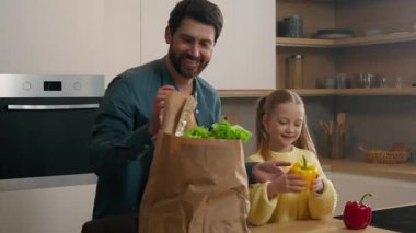 Mutfakta gülümseyen ebeveyn, mutlu beyaz çocuk kız çocuğu babasının sebze paketlerini açmasına yardım ediyor. Vitamin, sağlıklı beslenme ürünleri, biber, kırmızı biber, domates, ekmek.
