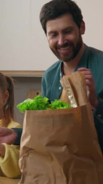 Mutfakta gülümseyen ebeveyn, mutlu beyaz çocuk kız çocuğu babasının sebze paketlerini açmasına yardım ediyor. Vitamin, sağlıklı beslenme ürünleri, biber, kırmızı biber, domates, ekmek.