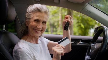 Neşeli, beyaz, yaşlı, yaşlı, gülümseyen, olgun bir kadın, otomobil şoförü, araba şoförü, araba anahtarı ve kredi kartını gösteren kameraya bakan, yeni bir araba kredisi satın alan,