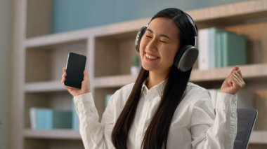 Japon iş kadını kadın işkadını Koreli kablosuz kulaklıklar takan Koreli işkadını cep telefonu uygulaması kullanıyor. Ofisteki müzik şarkısının akıllı telefon listesi sesini dinliyor.