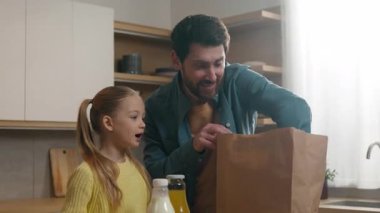 Evde eğlenen beyaz bir aile süpermarketten yiyecek getirdiler. Babaları, küçük kızları, küçük kızları, alışveriş çantalarını boşaltıyorlar. Elmalı domates ve doğal ürünler satan bir dükkan.