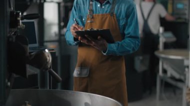 Küçük işletme sahibi Afrikalı Amerikalı erkek adam kavrulmuş kumaş kontrolünde çalışıyor kahve çekirdeklerini kavuruyor teknisyen kontrol makineleri kalite kontrolü yapıyor notlar yazıyor.