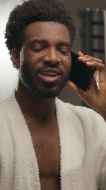 Bornozlu Afrikalı Amerikalı bir adam cep telefonuyla konuşuyor aynaya bakıyor gülümseyen bir etnik adam vücut bakımı sabah duş hijyenik sakallı bir erkek otel banyosunda akıllı telefon konuşması yapıyor.
