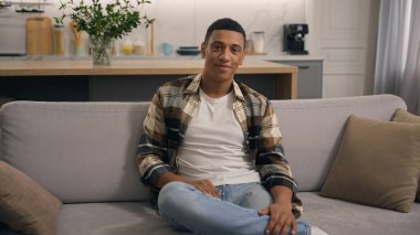 Evde gülümseyen, neşeli, samimi, genç, sakin, oturma odasında dinlenen Afro-Amerikan bir adam. Sadece haftasonu kanepede kameraya bakıyor.