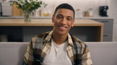 Evde gülümseyen, neşeli, samimi, genç, sakin, oturma odasında dinlenen Afro-Amerikan bir adam. Sadece haftasonu kanepede kameraya bakıyor.