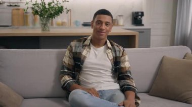 Evde gülümseyen, neşeli, samimi, genç, sakin, oturma odasında dinlenen Afro-Amerikan bir adam. Sadece haftasonu kanepede kameraya bakıyor.