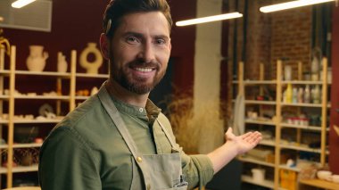 Neşeli gülümseyen beyaz adam, iş adamı, apron satıcısı, çömlekçi görünümlü küçük işletme sahibi, kameraman, işaret parmağıyla hoş geldin diyor.