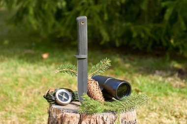 Siyah taktik bir bıçak, klasik bir monoküler, bir el bilgisayarı, ve yeşil çimenlerin bulanık arka planına karşı tahta bir kütüğün üzerinde dinlenen bir çam dalı..