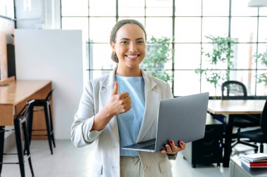 Güzel, kendine güvenen İspanyol ya da Brezilyalı bir kadın, yönetici, ofiste şık giysiler içinde duruyor, elinde açık bir dizüstü bilgisayar tutuyor, baş parmak işareti yapıyor, kameraya bakıyor, gülümsüyor.