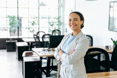 Başarılı Brezilyalı ya da İspanyol iş kadını, şirket çalışanı, yönetici, ceketli, kollarını ofisin arka planında kavuşturmuş, kameraya bakıyor, gülümsüyor.