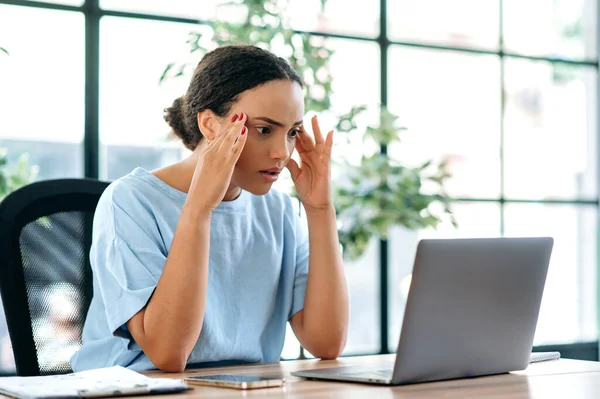 Surprised frustrated mixed race office worker, sitting at a desk in the office, looking disappointed at the laptop screen, reading bad news, bad work result, experiencing negative emotions