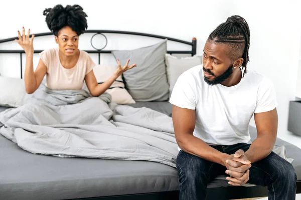 stock image Relationship problems, disagreements. Upset african american husband sits on bed, his wife scolds him, screams emotionally and gesticulates with hands. Quarrel between spouses, conflict