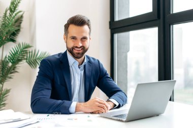 Takım elbiseli yakışıklı, kendine güvenen başarılı beyaz bir adamın portresi, IT uzmanı, yatırımcı, CEO, modern bir ofiste bir iş yerinde oturur, kameraya bakar, dostça gülümser.