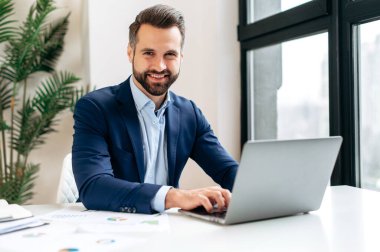Bir iş adamının portresi. Çekici, kendine güvenen, takım elbiseli başarılı beyaz adam, bilişim uzmanı, yatırımcı, CEO, modern bir ofiste iş yerinde oturuyor, kameraya bakıyor, gülümsüyor.
