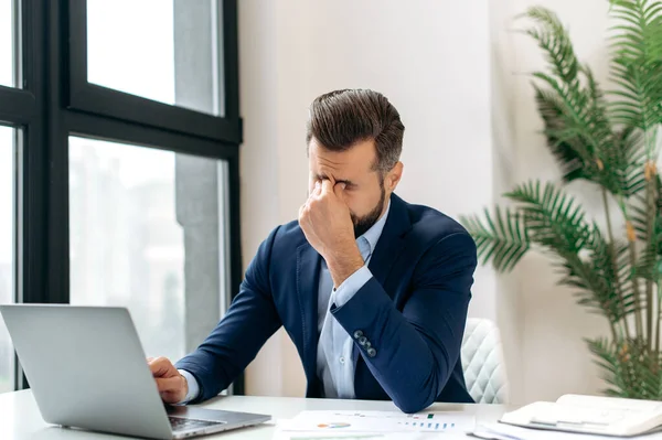 Overworking. Frustrated tired caucasian entrepreneur, product manager, programmer, sit at a desk in the office, massaging the bridge of his nose with his eyes closed, feeling exhausted from long work
