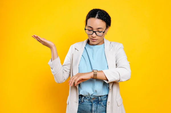 Confused mixed race young woman, company employee, using smart watch on her hand, looks at the time, worried through be late, waiting for someone, stands on isolated orange background
