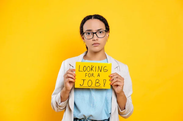 忧心忡忡的男女混血的年轻女子戴着眼镜 富有创意地寻找工作 站在孤立的橙色背景上 带着寻找工作的迹象 失望地看着镜头 — 图库照片