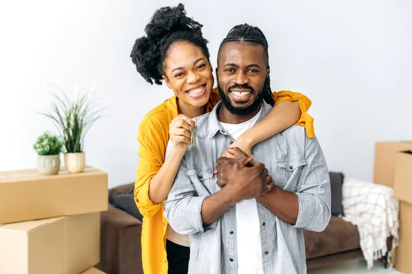 New housing, relocation. Happy joyful african american family, black couple hugging while standing in living room, woman showing keys of their new property, they looking at camera, smiling