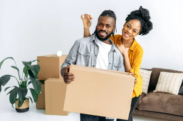 幸福的丈夫和妻子 站在新客厅里 男人拿着大盒子 女人拿着钥匙 他们看着相机 有爱心的非洲裔美国夫妇搬进他们的新家 — 图库照片