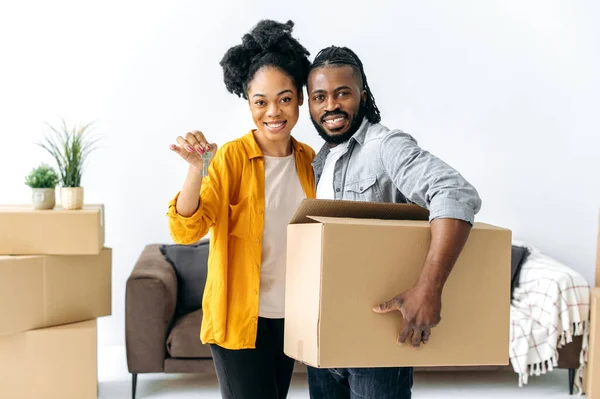 有爱心的非洲裔美国夫妇搬进了他们的新家 幸福的丈夫和妻子 站在新客厅里 男人拿着大盒子 女人拿着钥匙 他们看着相机 微笑着 — 图库照片