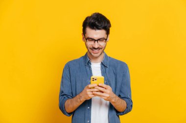 Yakışıklı beyaz adam akıllı telefonunu kullanıyor, arkadaşlarıyla mesajlaşıyor, internette ve sosyal ağlarda geziniyor, izole edilmiş turuncu bir arka planda duruyor, gülümsüyor.