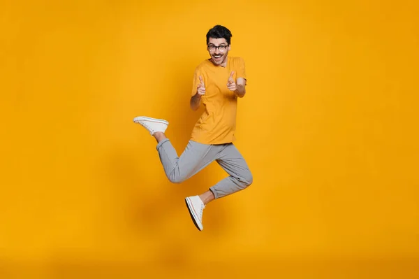 Full length headshot of a cheerful funny caucasian smart guy with eyeglasses, stylishly dressed, smiling and jumping high running on isolated orange background, gesturing hands
