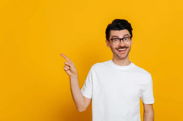stock image Excited caucasian happy guy in white basic t-shirt and glasses amazed looks at camera and points finger to the side at space for your presentation, stands on isolated orange color background, smiling