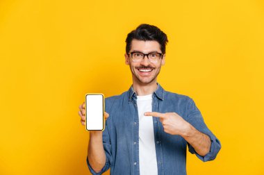 Heyecanlı, mutlu, tıraşsız, kot gömlekli ve gözlüklü beyaz adam mesajın için beyaz ekranlı akıllı telefonu tutuyor ve onu işaret ediyor izole edilmiş turuncu arka planda duruyor, gülümsüyor.