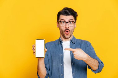 Şaşırdım. Kot gömlekli ve gözlüklü beyaz adam mesajın için beyaz ekranlı akıllı telefonu tutuyor ve onu işaret ediyor. İzole edilmiş turuncu arka planda duruyor.