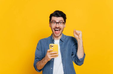 Neşeli, heyecanlı, modern, pozitif beyaz adam elinde akıllı telefon tutuyor, arkadaşlarıyla mesajlaşıyor, internette, sosyal ağlarda geziniyor, beklenmedik haberler alıyor, kazanıyor, izole edilmiş turuncu arka planda duruyor, gülümsüyor.
