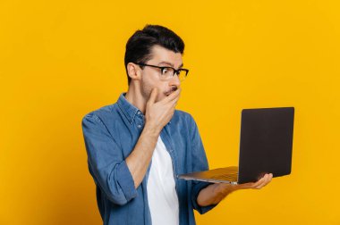 Kafası karışmış, kafası karışmış, kafası karışmamış, kafkasyalı gözlüklü, elinde bir dizüstü bilgisayar tutan adam şaşırmış görünüyor. Ağzı ağızla kaplarken, izole edilmiş turuncu arka planda duruyor.