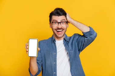 Şaşırdım. Kot gömlekli ve gözlüklü beyaz adam mesajın için beyaz ekranlı akıllı telefonu tutuyor. Kafaya elle dokunuyor, izole edilmiş turuncu arka planda duruyor.