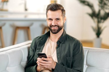 Mutlu, yakışıklı, beyaz sakallı bir adam, evinde bir kanepede oturuyor, akıllı telefonunu kullanıyor, kanepede dinleniyor, sosyal medyada mesajlaşıyor, internette geziniyor, kameraya bakıyor, gülümsüyor.