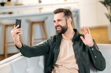 Arkadaş canlısı, yakışıklı beyaz adam, şık gündelik giysiler içinde, evde rahat bir koltukta otururken arkadaşlarıyla ya da ailesiyle online video konferansı yapıyor, el sallıyor, gülümsüyor.