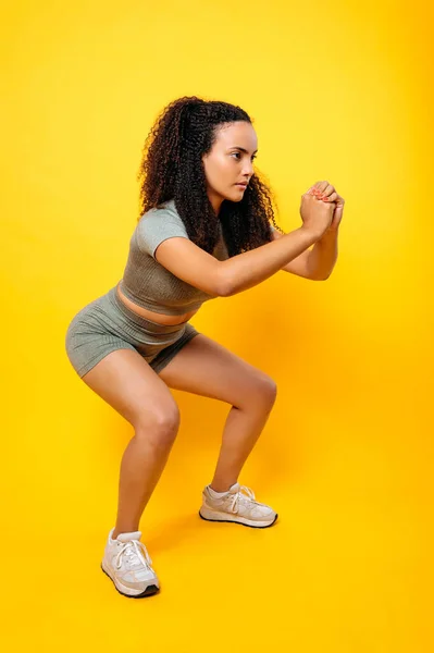 Sports lifestyle. Full length vertical photo of a fitness sporty lovely curly haired brazilian or latino woman in sportswear, practicing squat exercise on isolated orange background