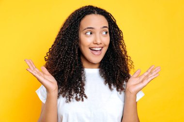 Surprised amazed cheerful positive brazilian or hispanic young woman with curly hair, looking away spreading her arms to the sides, standing over isolated yellow background