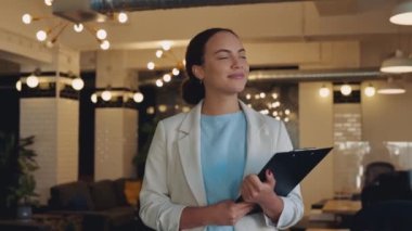 Positive confident successful brazilian or hispanic business woman, wearing stylish clothes, go across the creative office, holding documents in her hands, looking at the camera, smiling