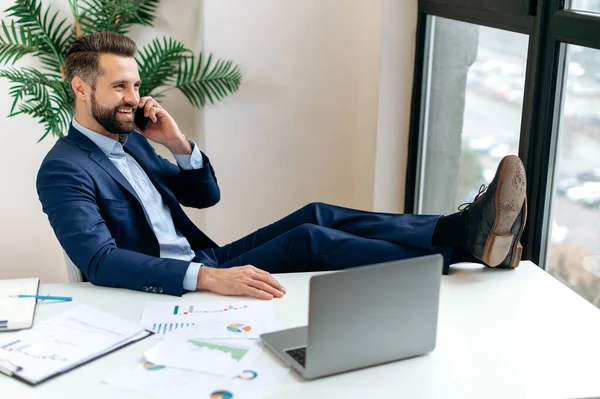Happy elegant caucasian business man, manager, director, sits at a desk in the office, talking on the phone with a colleague or client, putting his legs on the table, making an appointment, smiling