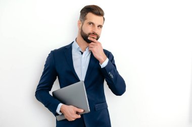 Successful confident caucasian businessman in a suit, seo, executive, broker, financial adviser, standing on isolated white background, holding laptop, looking to the side, thinking over idea,dreaming