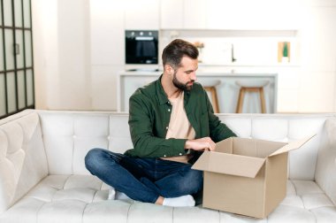 Excited caucasian stylish man in casual wear, unpacks his long awaited package, examines the clothes, happy with a good purchase, sits at home on the sofa in the living room. Online shopping concept