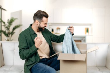 Cheerful stylish caucasian man opening big carton box while sits on sofa, unpacking internet store order at home, feel satisfied with fast delivery service and the goods which received, YES gesture