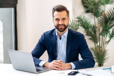 Photo of a handsome confident successful positive businessman, financial director, entrepreneur, dressed in an elegant suit, sitting at a work desk in the office, looking at the camera, smiling