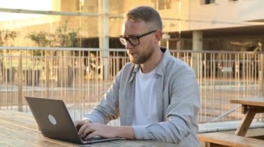 Smart confident successful caucasian bearded man with glasses, programmer, seo, sits at a desk with a laptop outdoors, drink a cup of coffee, working online on a project, develop strategy